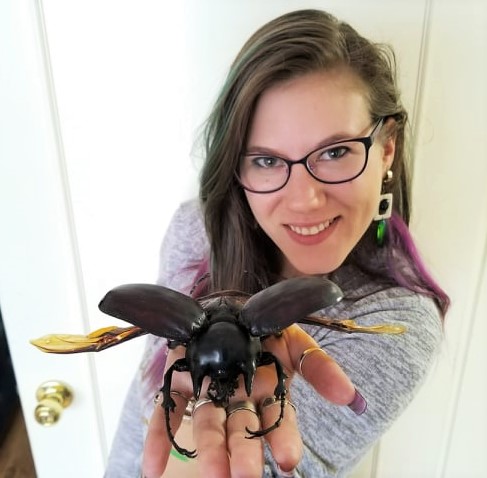 A long haired woman with purple glasses. Her hand is outstretched and in it is a large, black,  horned beetle taking up her entire hand. 