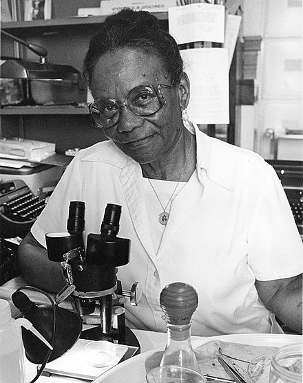 An African American woman sitting in a white lab coat in front a microscope