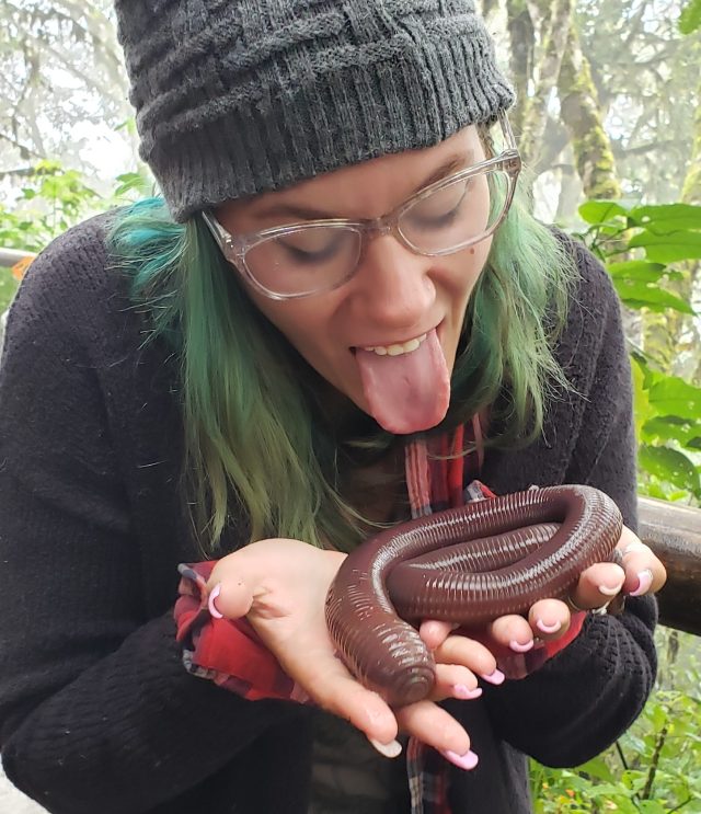 A girl in a beanie with green hair, arcrylic nails holding a giant earthworm measuring about 3ft in length and shes about to lick it